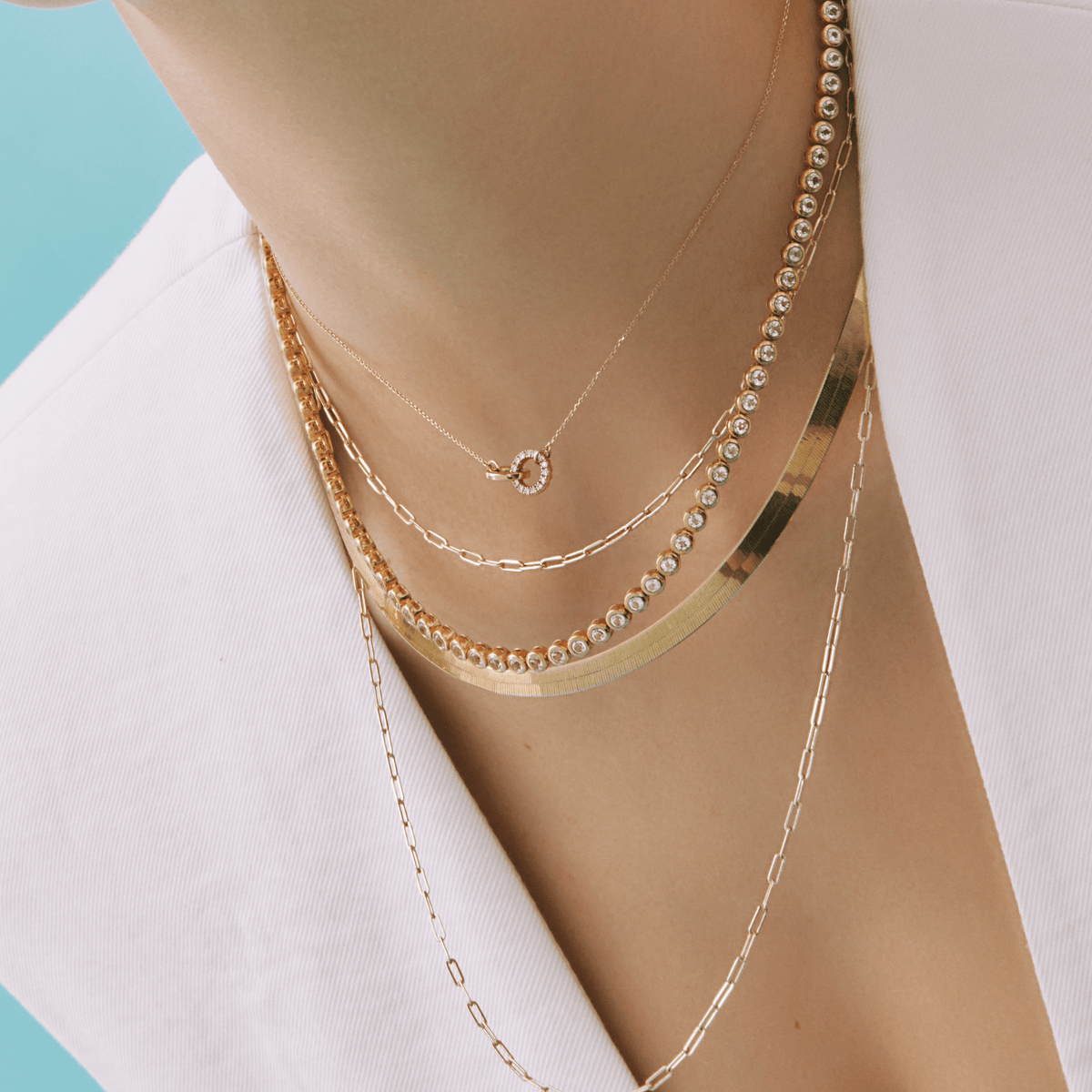 Lifestyle Model Close-up of a woman wearing a white top and five layered gold necklaces, including a delicate chain with a circular interlocking pendant, a paperclip chain, a diamond tennis necklace, a gold herringbone chain, and a long delicate chain.