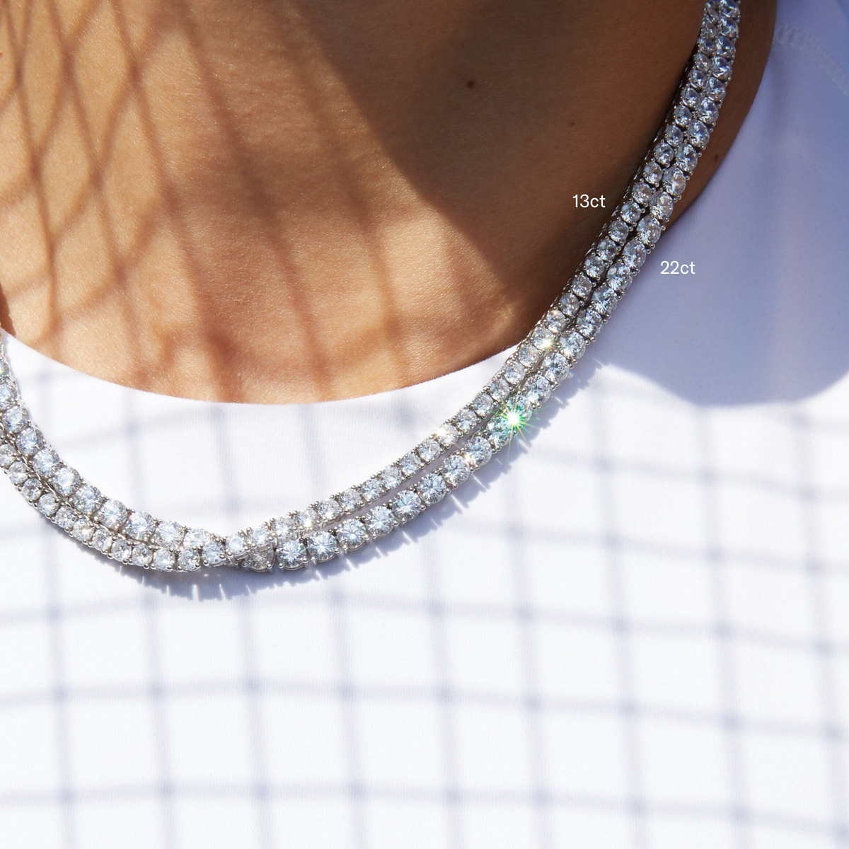 Model. Close-up of a woman's neck and collarbone, wearing a white top and two layered white sapphire tennis necklaces. A distinctive lattice shadow falls across her skin, and text overlays label the top necklace as '13ct' and the bottom necklace as '22ct'