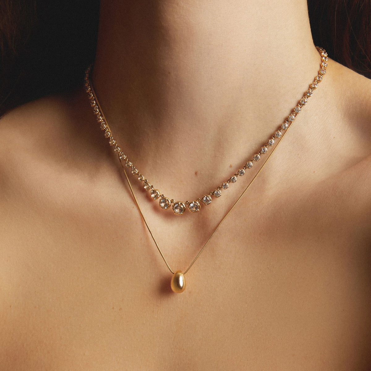Model. One diamond tennis necklace and one gold pendant necklace stacked at different lengths on a woman.