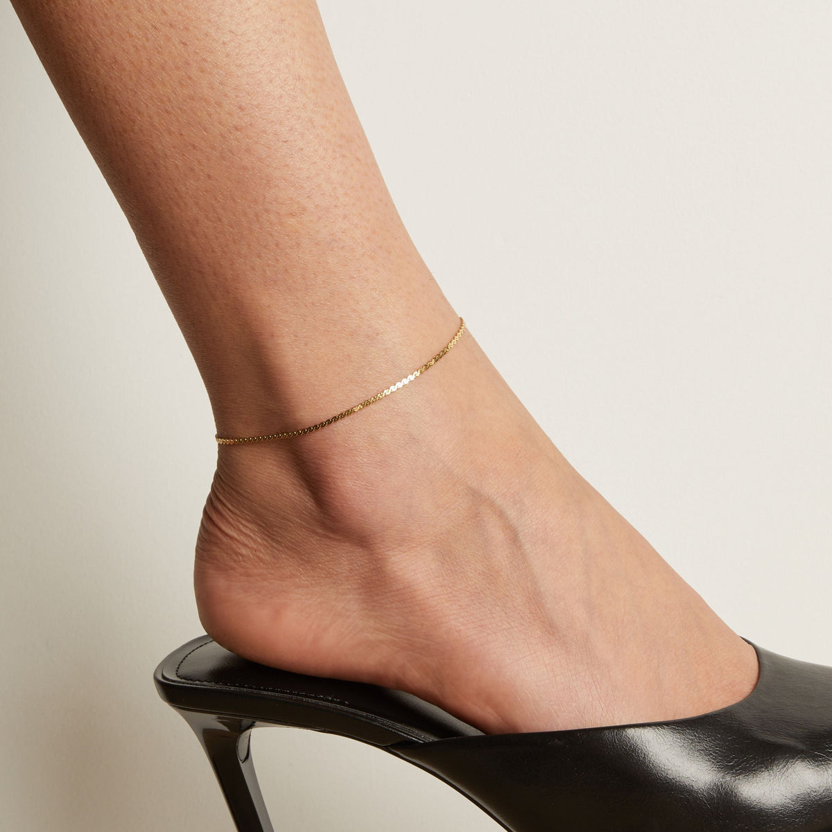 Model. Gold anklet on a foot wearing a black high heeled shoe against a neutral background.