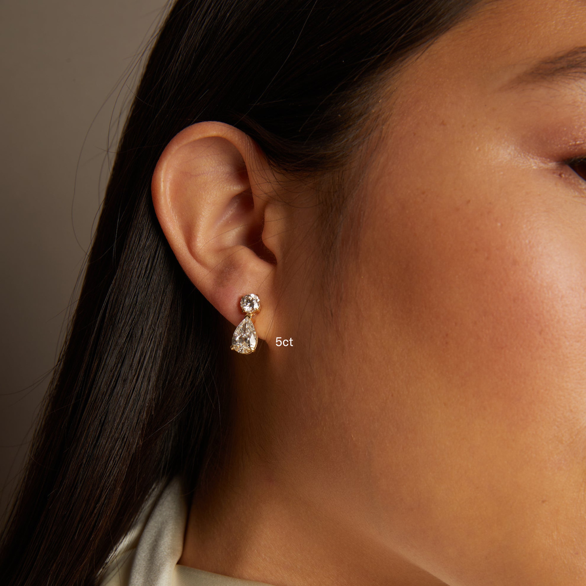 Model. Close-up of an ear wearing a diamond earring with '5ct' label against a neutral background