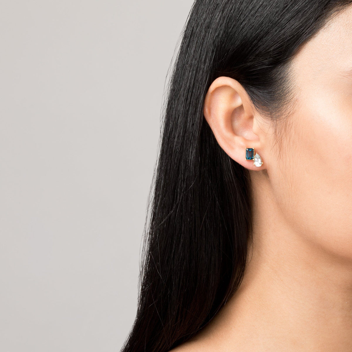 Model Close-up of a woman wearing a gold two-stone earring with a blue and a clear gemstone.