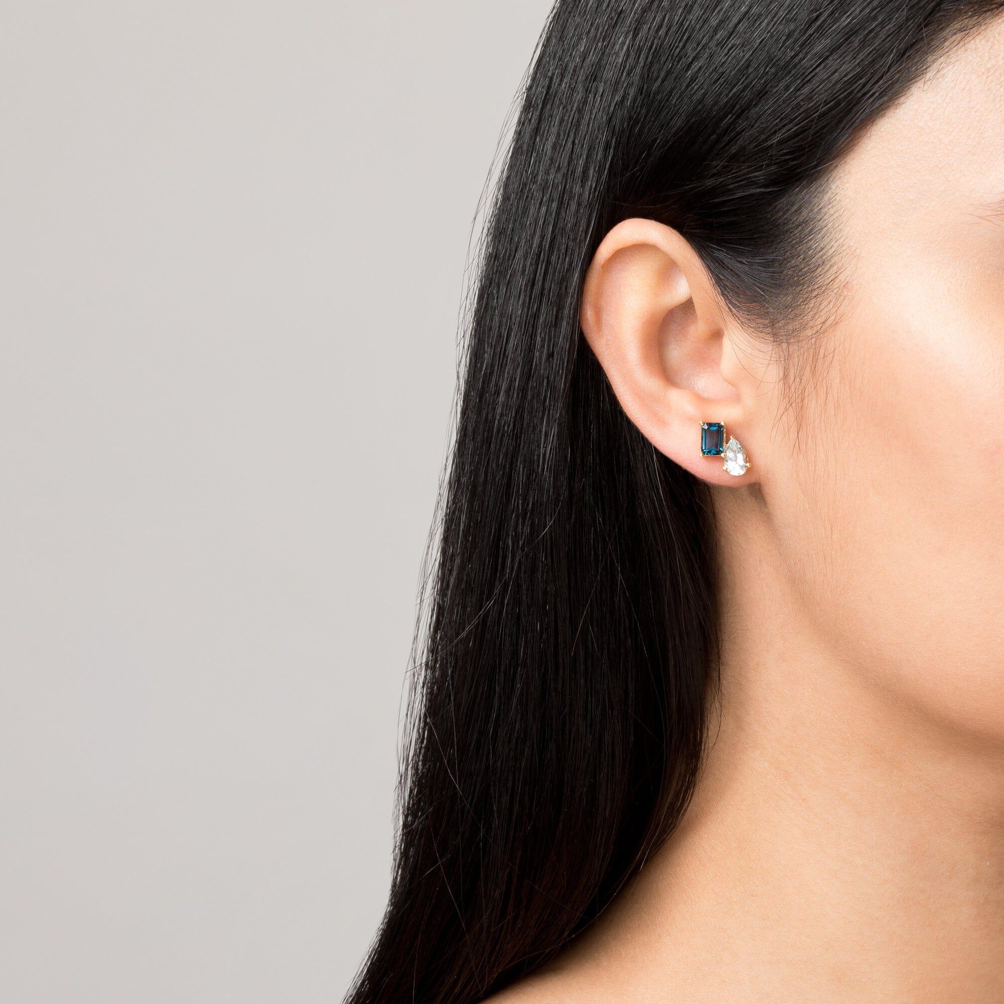 Model Close-up of a woman wearing a gold two-stone earring with a blue and a clear gemstone.