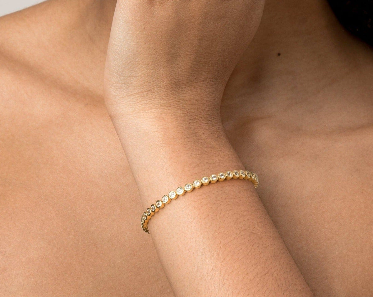 Model Close-up of a woman's wrist resting against her chest, wearing a single gold diamond bezel tennis bracelet.