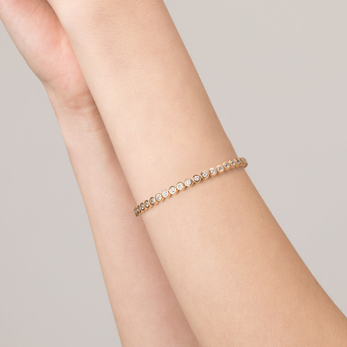 Model Close-up of a woman's raised arms wearing a single gold diamond bezel tennis bracelet against a light beige background.