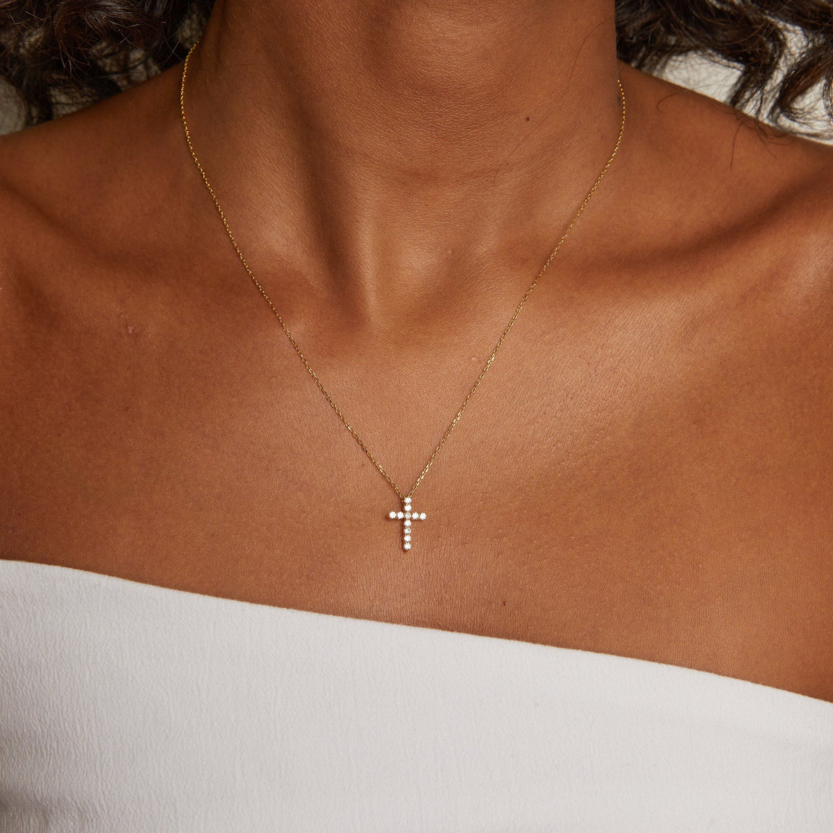 Model. Gold necklace with a cross pendant on a person wearing a white top.