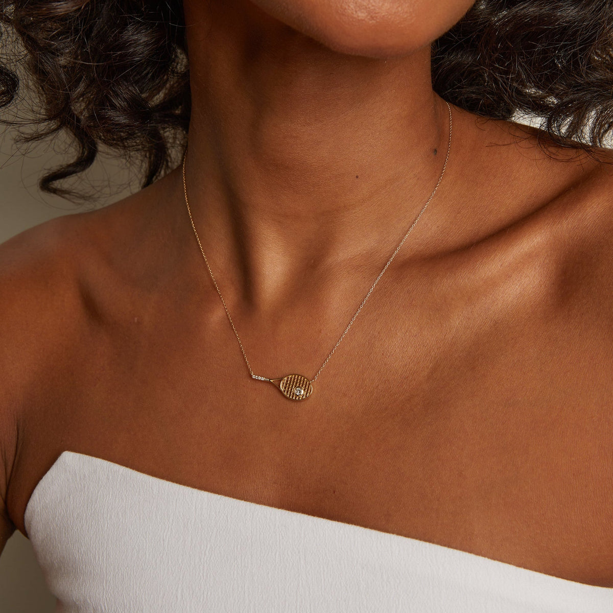 Model. Close-up of a woman wearing a gold necklace with a white top against a neutral background